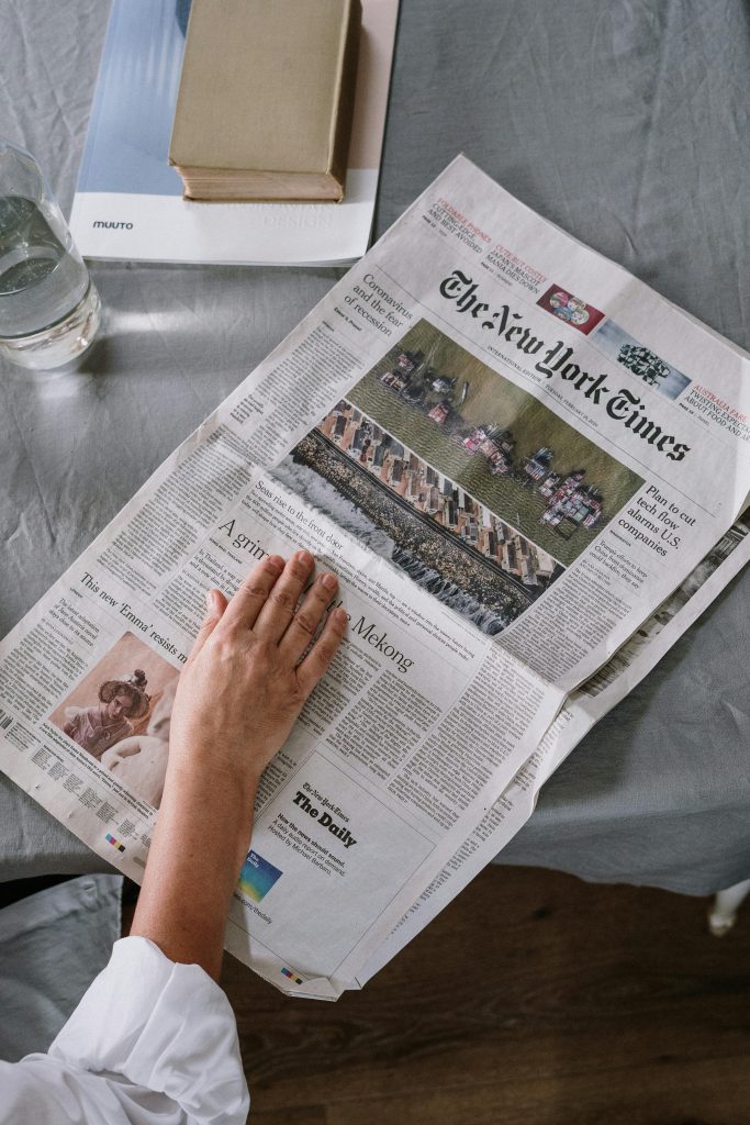 Person reading The New York Times at home with books and glass of water on the table.